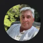 A man with gray hair wearing a white shirt, sitting outdoors with a neutral expression.